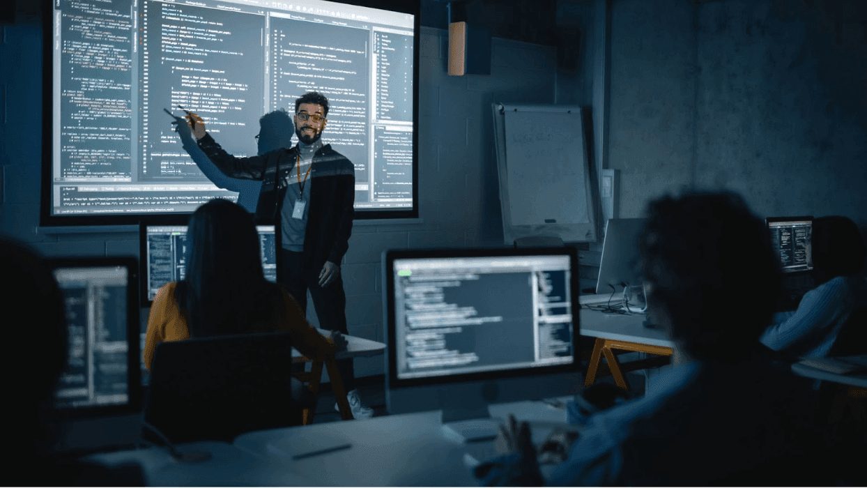 Teacher stands in front of a projector screen while demonstrating how to code in a computer class.
